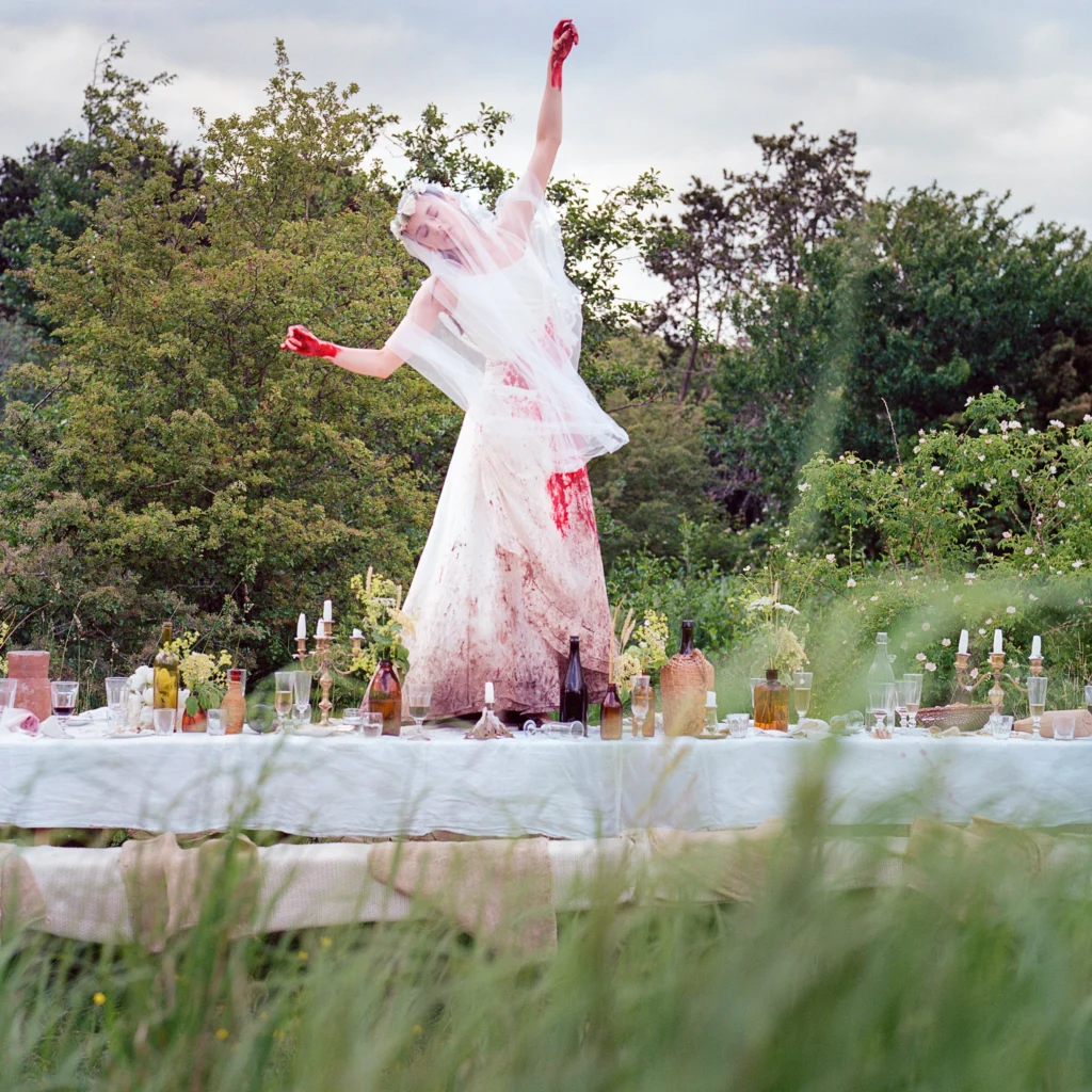 A woman in a blood-spattered wedding dress, her hands bathed in red, stands atop a long table set for a feast in the midst of a clearing in a forest.