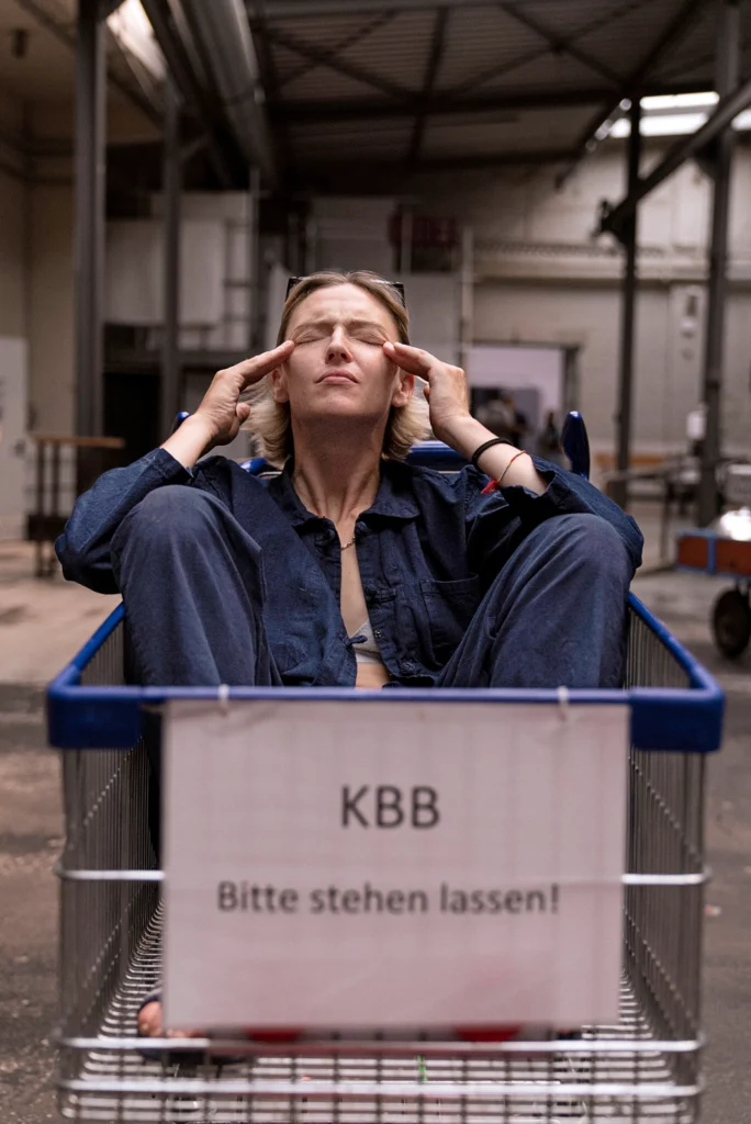 Oona Doherty sits in a shopping basket adorned with a sign that reads "KBB: Bitte stehen lassen!" Her fingertips rest against her temples as she screws her eyes shut and tips her chin up. She wears an unbuttoned navy blue jumpsuit.