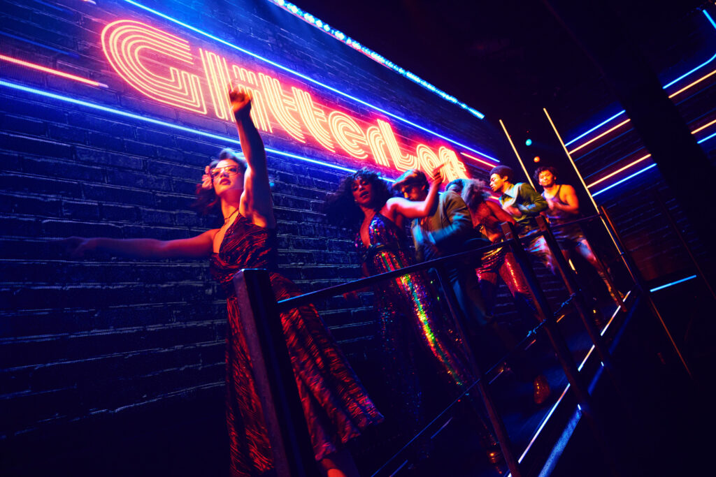 Six performers in disco finery dance down an overhead catwalk. "GlitterLoft" is spelled out above them in neon lights.