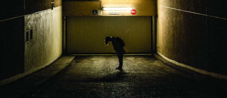 A figure stands silhouetted in the dull, greenish light of a parking garage. They lean forward, hair falling around their face as they look toward their shoes. They wear a backpack.