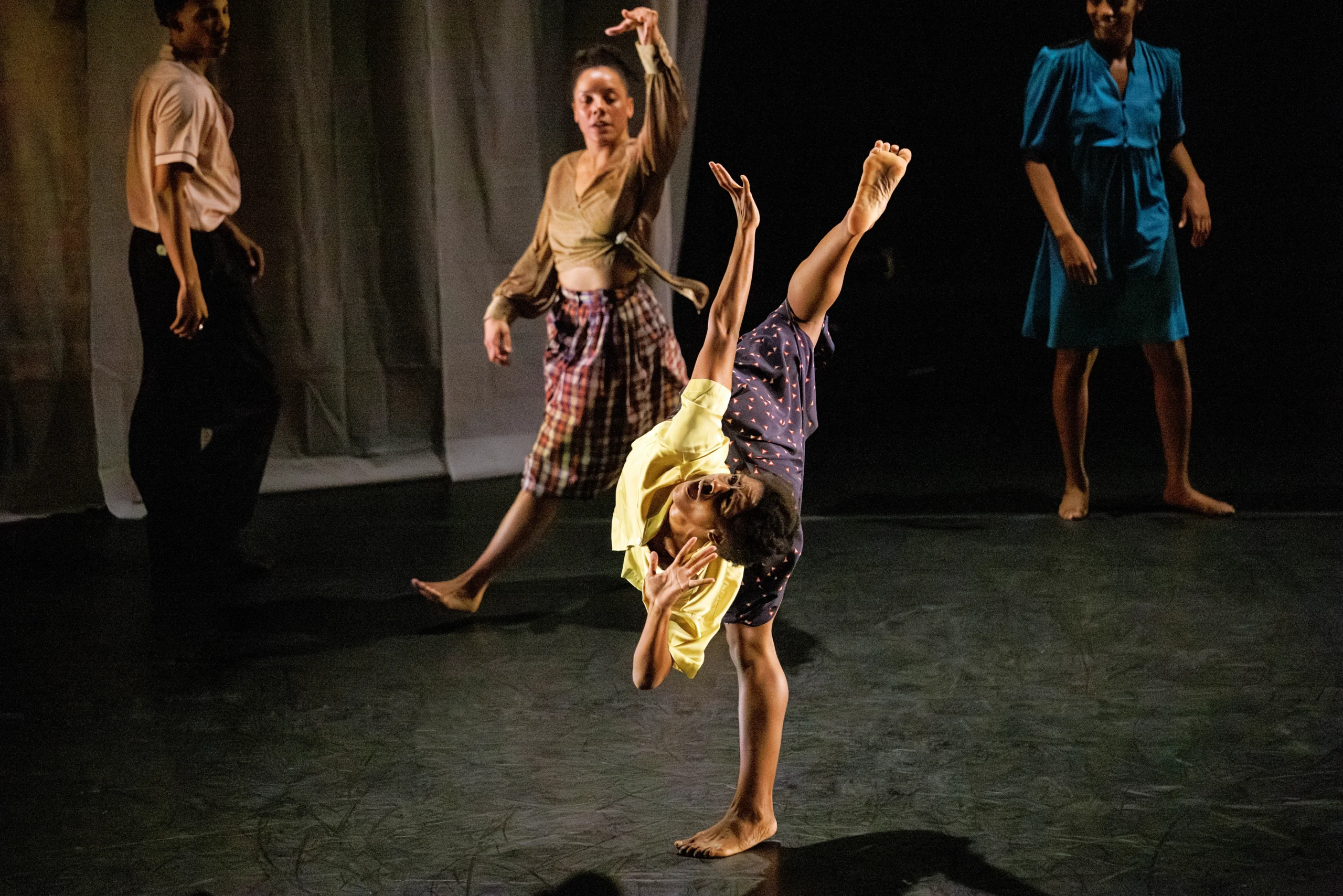 Kayla Farrish tips into an attitude penché, twisting her torso so it's parallel to her standing leg. One hand reaches up, the other is cupped by her mouth as though to amplify a shout. She wears a yellow blouse and an indigo polk-dotted skirt. Three dancers walk past in the background, faces turned toward her.