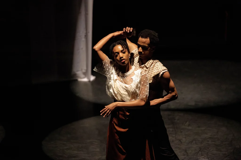 A female dancer stands just in front of her partner, raising one hand overhead as he turns her slowly in place. Her expression is thoughtful, or dreamy. She wears a white polka-dotted blouse.