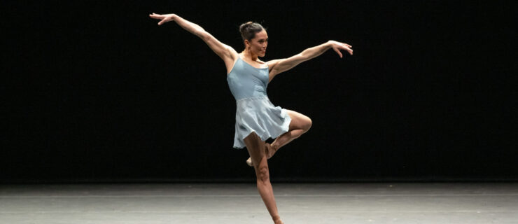 Lia Cirio on stage wearing a light blue leotard and skirt poised in a retire with her arms extended side