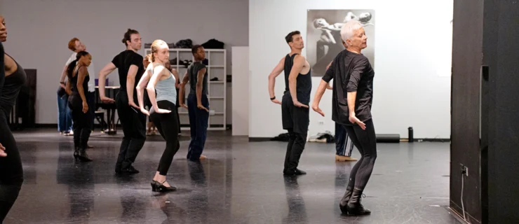 A studio of dancers imitate a signature Fosse position, legs in parallel and knees bent, arms lightly flexed behind them as they look over their right shoulder.