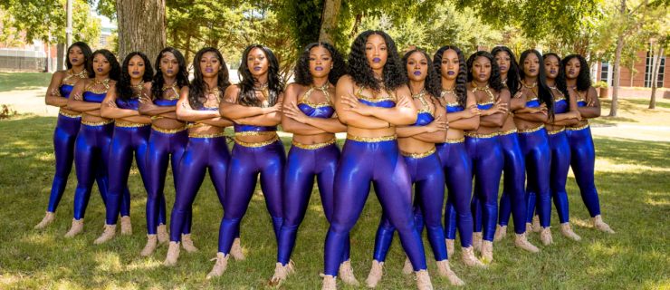 a group of Black women standing with their arms crossed in a V formation. They're wearing purple and gold uniforms with dark lipstick