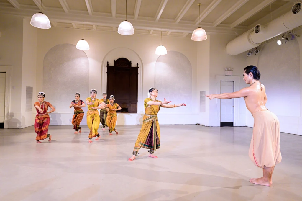 A woman reaches with open hands toward a figure in a nude shift. In the background, five dancers step one foot over the other, hands making the same gesture at waist height.