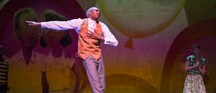 A dancer with silver hair, wearing an old fashioned vest over a button down and gray pants, presses one hand to his heart. His gaze is distant as he rolls through relevé, one arm stretched beside him in opposition as if for balance. In the background, a woman in a dress watches, smiling, with her hands clasped.