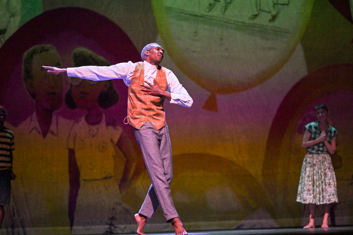 A dancer with silver hair, wearing an old fashioned vest over a button down and gray pants, presses one hand to his heart. His gaze is distant as he rolls through relevé, one arm stretched beside him in opposition as if for balance. In the background, a woman in a dress watches, smiling, with her hands clasped.