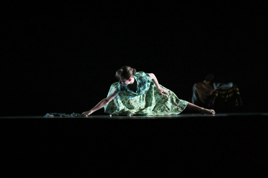 In shadowy lighting, a woman in a green patterned dress lunges to the floor to drag a piece of fabric against the floor.