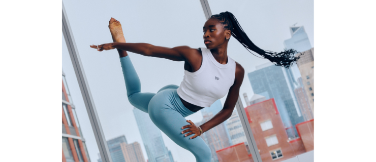 Caroline T. Dartey dancing in a studio with large windows overlooking the city. She is wearing blue leggings and a white crop top.