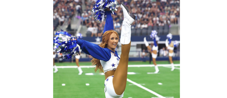 Charly Barby high kicking on the field wearing a blue and white uniform while holding poms