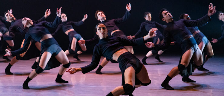 Eight dancers hinge back toward the floor, arms open but drooping. The most downstage dancer turns their head to look at the audience. They wear black socks, kneepads, trunks, and cropped long sleeve shirts.