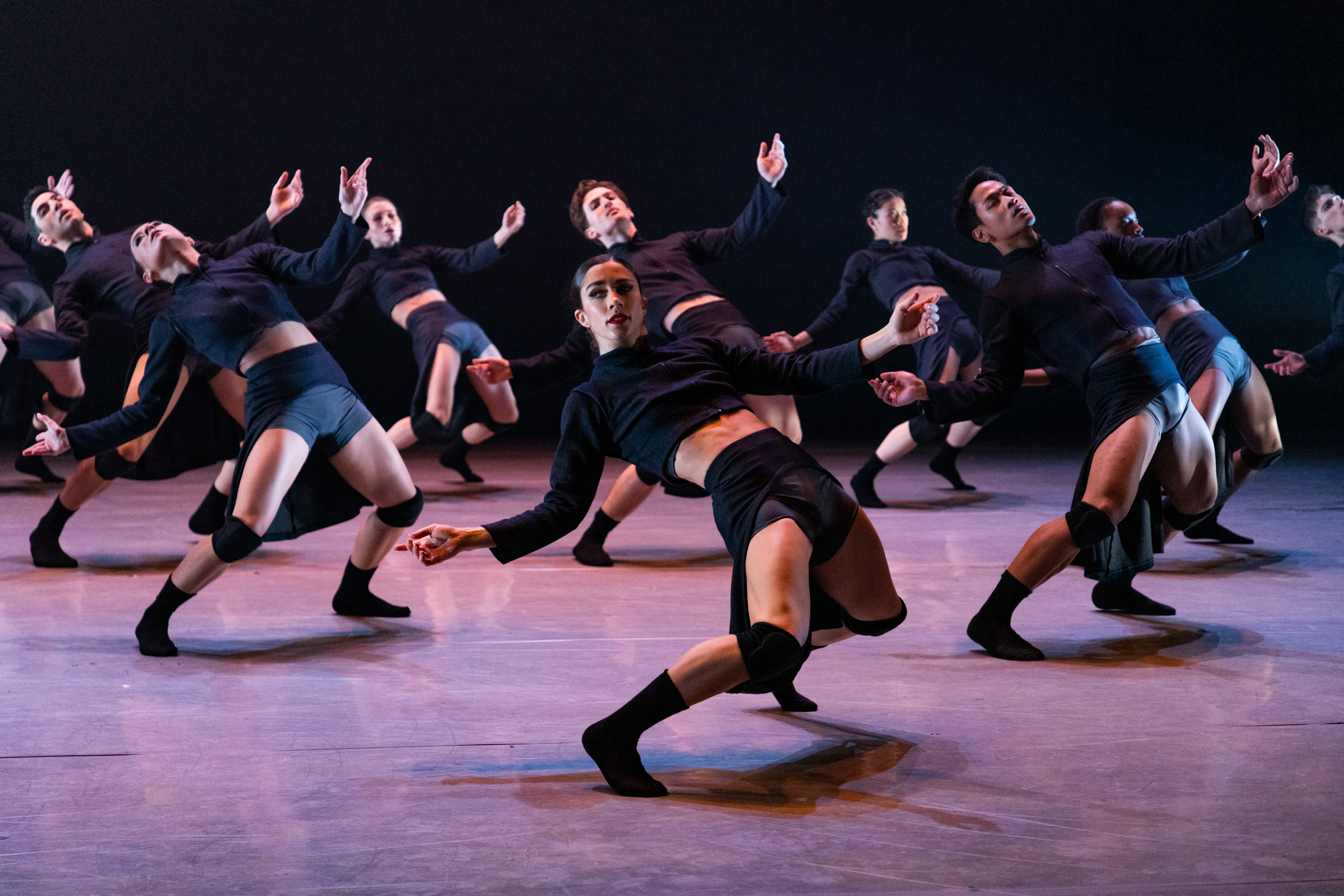 Eight dancers hinge back toward the floor, arms open but drooping. The most downstage dancer turns their head to look at the audience. They wear black socks, kneepads, trunks, and cropped long sleeve shirts.