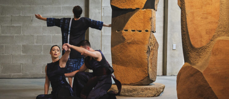 3 dancers performing in the Noguchi Museum. They dancer next to a large stone sculpture wearing dark costumes