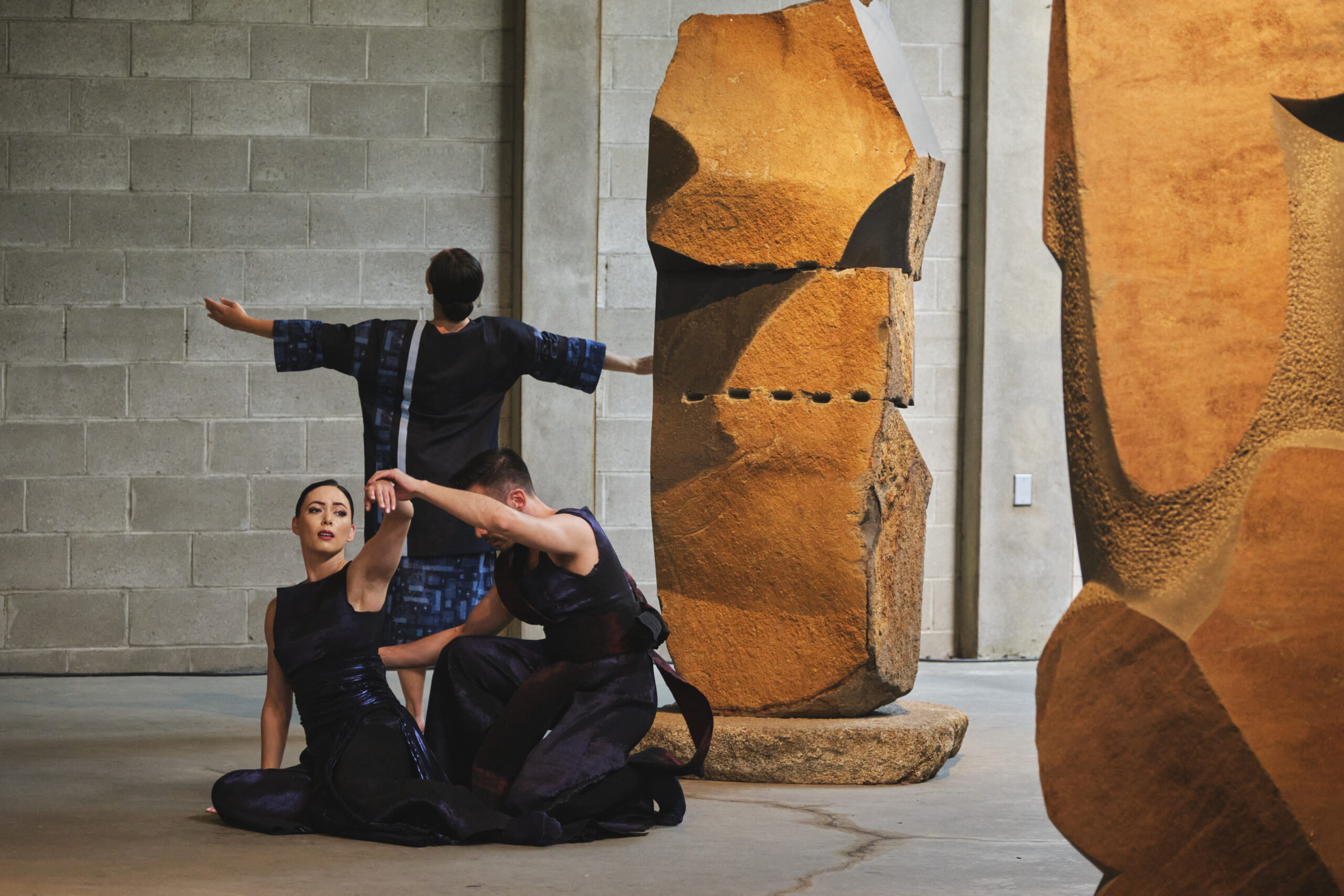 3 dancers performing in the Noguchi Museum. They dancer next to a large stone sculpture wearing dark costumes