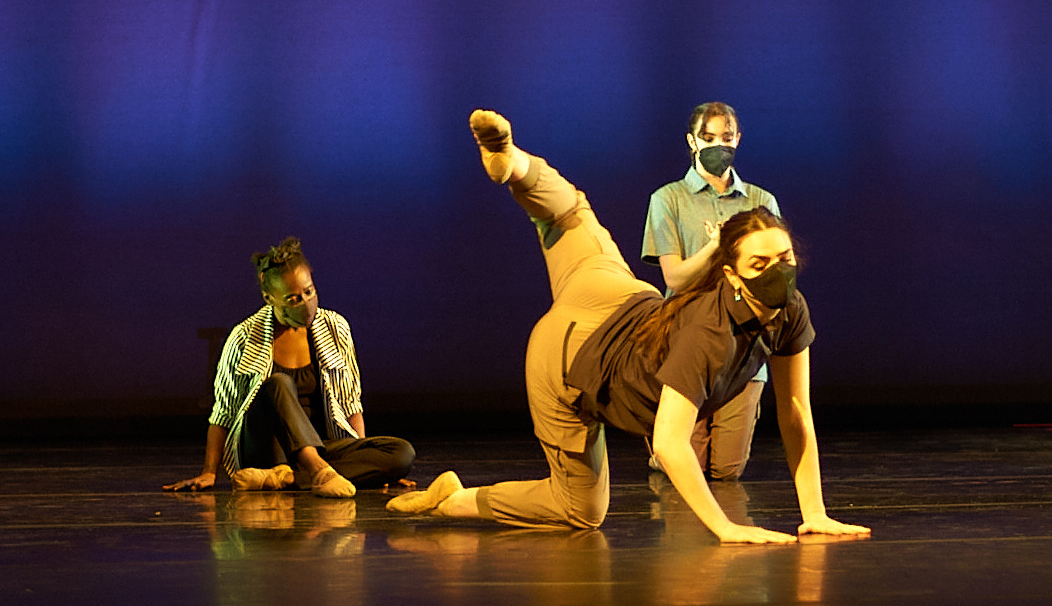 Three dancers wearing loose neutral-colored shirts and pants and black face masks are pictured in a triangle formation on stage. The dancer in front supports herself on her hands and her right knee as she extends her left leg behind her in attitude. The dancer at back left sits with her hands braced behind her and her right leg crossed over her left. The dancer at back left kneels with one hand on her chest.