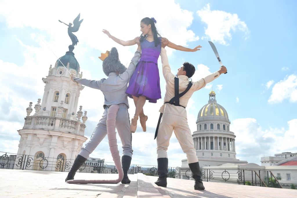 A trio of dancers on a rooftop surrounded by impressive buildings. The Rat King and Nutcracker Prince each support Clara under one arm as they lift her overhead; their outside hands hold swords. Clara's arms are outstretched to either side as she looks down at the Rat King in alarm. She wears a vibrant purple dress and pointe shoes.