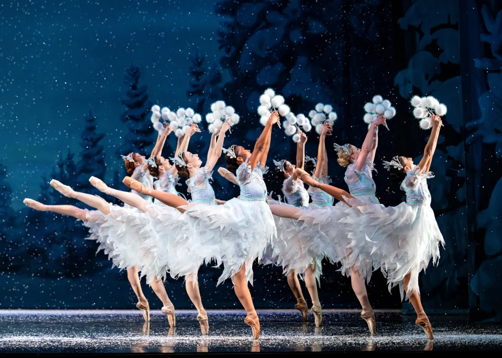 Two rows of four ballerinas balance in first arabesque on pointe, clusters of snowflakes raised overhead. The backdrop is of a snowy wood at dusk; fake snow falls to the stage around them.