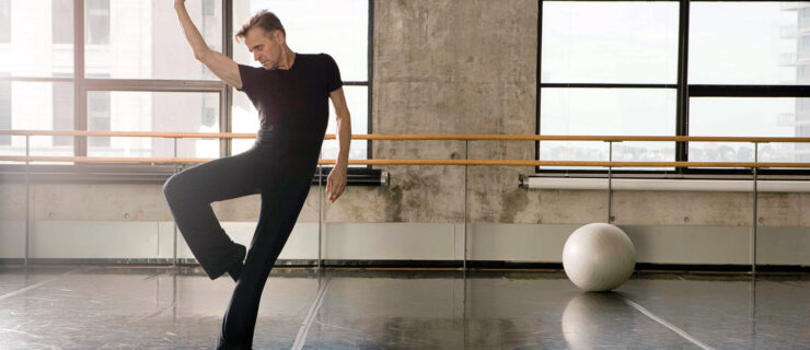 Baryshnikov–a middle-aged white man wearing black dance clothes—poses in retiré, with his right arm elegantly framing his face, in a sunlit industrial dance studio.