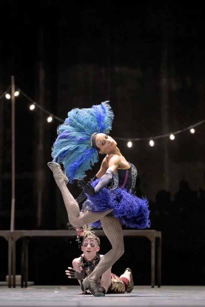 Leroy Mokgatle looks over her shoulder toward the camera, bent back leg kicking up toward the back of her head as she pushes a hand toward the camera. She wears a blue feathered headdress that matches a feathery short skirt, fishnets, heels, and elbow length black gloves. A dancer upstage lies on his stomach and props up on his elbows to clap.