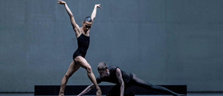 Leroy Mokgatle in performance. She poses with her front leg in forced arch on pointe, arms in a V slightly below her head, head turned down to watch her partner sweep the floor around her feet. She wears a black leotard and pointe shoes pancaked to match her skin tone. Her partner is dressed in sheer black.