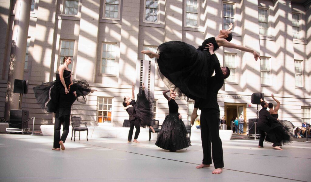 Dana Tai Soon Burgess Dance Company dancers wearing black costumes, long tutus for the women and pants for the men, as they lift each other around the stage. They perform outside in front of the National Portrait Gallery 