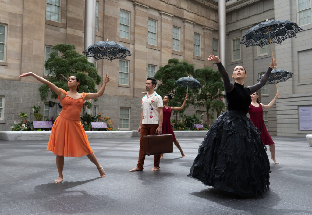 Dana Tai Soon Burgess Dance Company dancers wearing a variety of costumes while dancing with parasols and a brief case. They perform outside in front of the National Portrait Gallery 