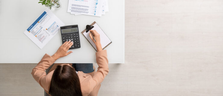 Overview of young female accountant with calculator sitting by desk, making calculations and writing down information on paper