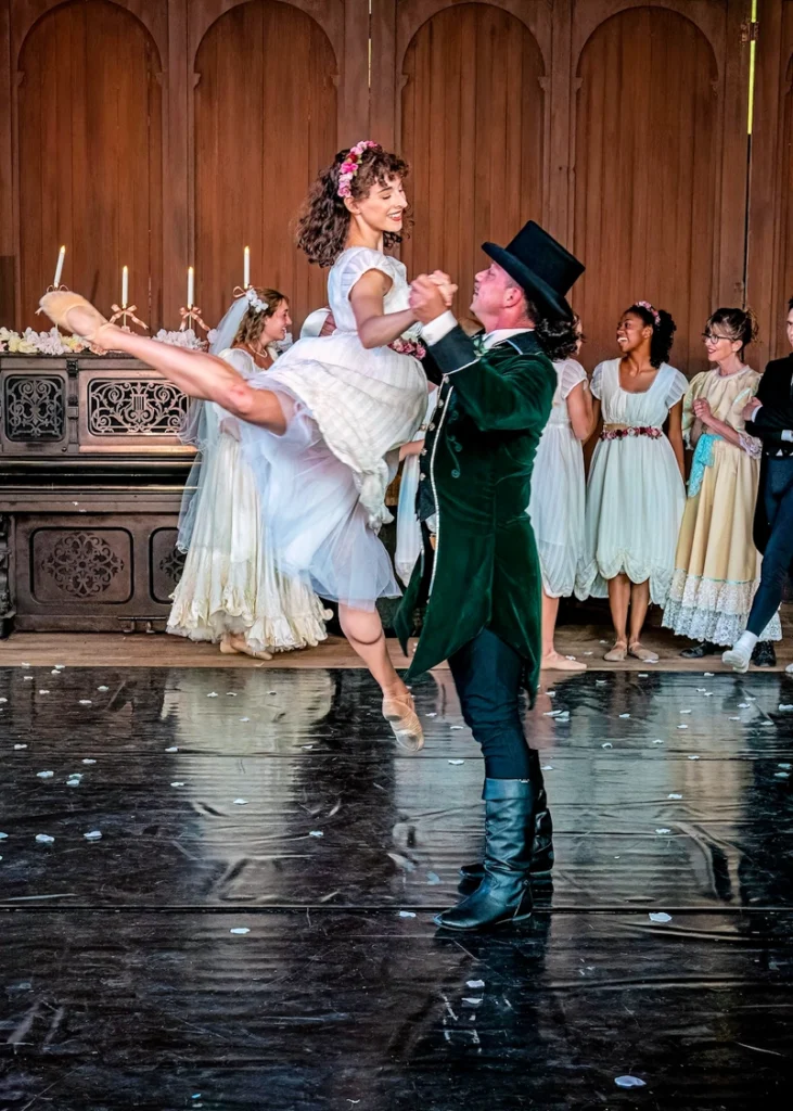 Jo March (played by AnnaLee Rohovec), in her beautiful cream bridesmaid attire and floral headpiece, is being lifted by Fredrich Bhaer (played by Jacob Robleto) in his emerald green wedding attire. The photo is taken at the Little Women Ballet Spring Immersive Experience in a duet between Fredrich and Jo during Meg's wedding that captures Jo's strong-willed personality.