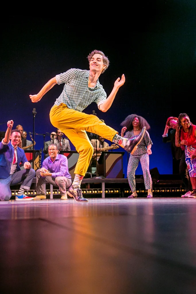 A dancer in bright yellow pants and a checkered button down smiles easily as he kicks a foot forward, arms working in opposition. He leans into the movement and looks over his shoulder downstage. Behind him, other dancers stand and crouch in a semicircle, clapping and cheering him on.