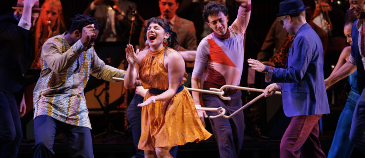 A cluster of dancers laugh and move together as a band plays upstage. Most prominent is a woman in a bright yellow dress, throwing her head back and laughing as she claps to the beat.