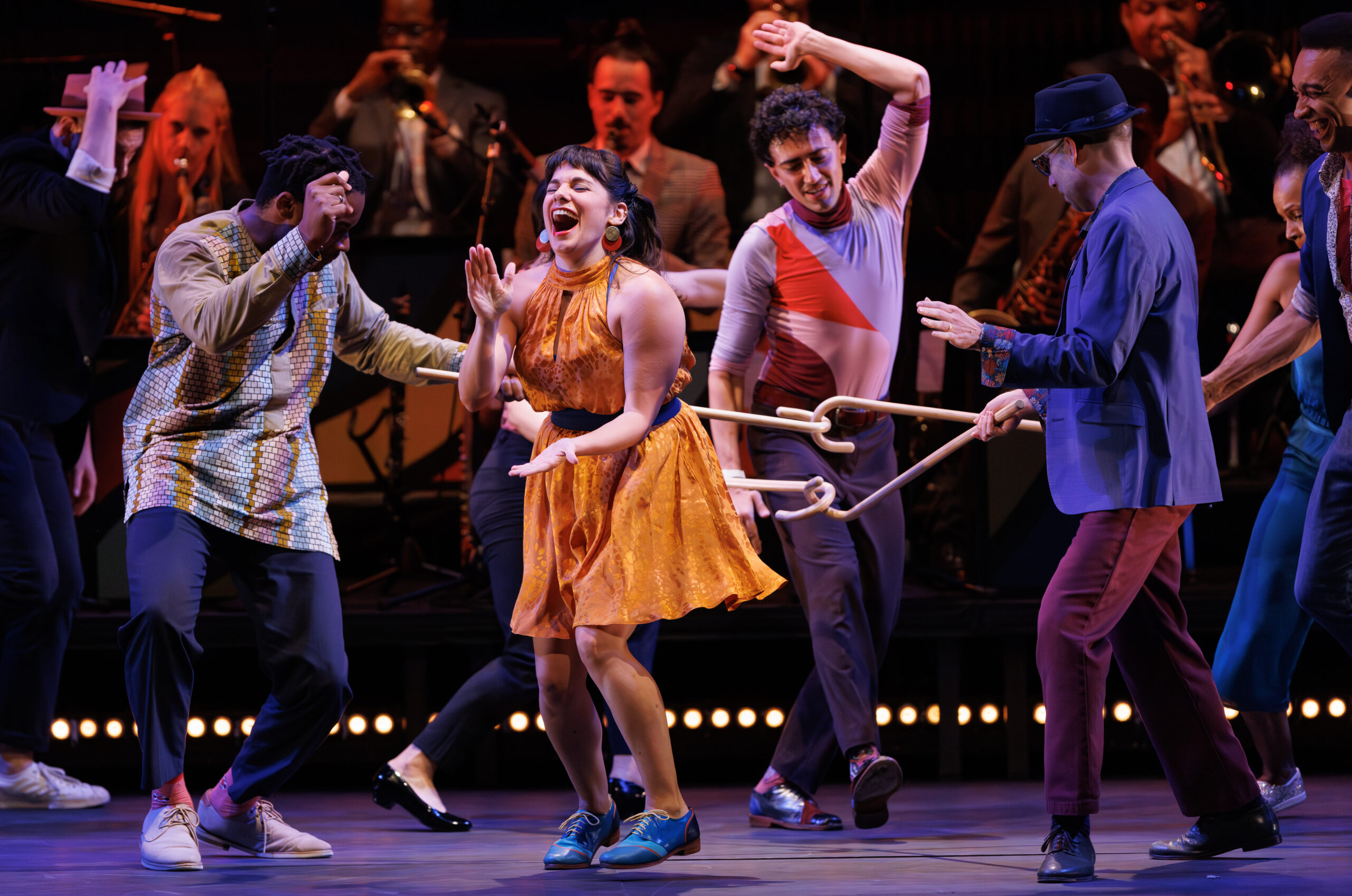 A cluster of dancers laugh and move together as a band plays upstage. Most prominent is a woman in a bright yellow dress, throwing her head back and laughing as she claps to the beat.