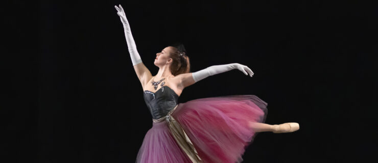 Laine Habony on stage in a purple romantic tutu. She performs a first arabesque en pointe