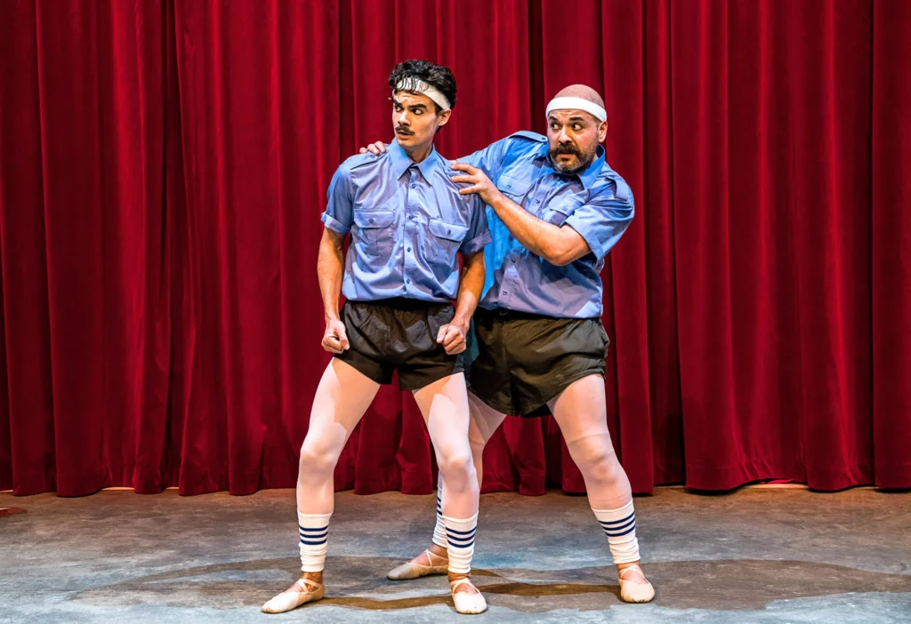 Two performers stand in front of a lush red theater curtain, looking nervously off to the corner, one hiding behind the other with hands on his shoulders. They are costumed identically—white sweatbands, blue button downs, black gym shorts over pink tights, white legwarmers, and ballet slippers.