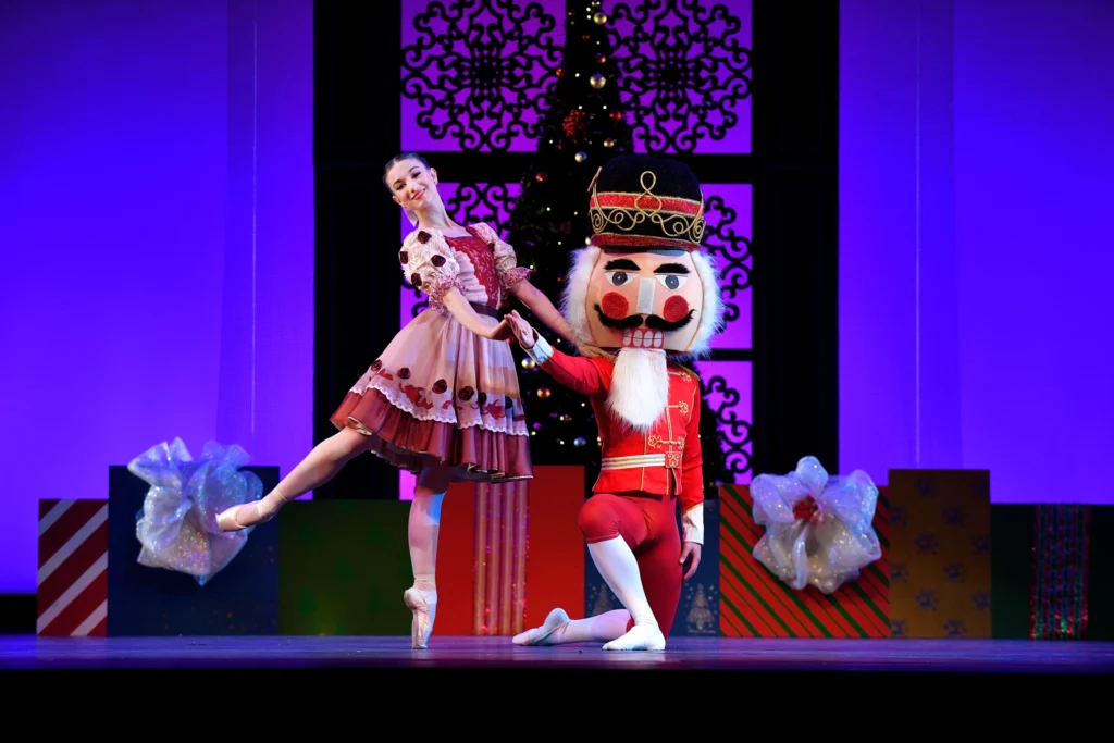 In front of a set evoking presents in front of a Christmas tree, the Nutcracker Prince kneels and offers a hand to Clara, who takes it and balances in a low back attitude on pointe.