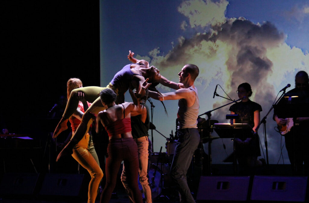 Owen/Cox Dance Group artists lifting a stretched out dancer in the air. Live musicians stand in the background 