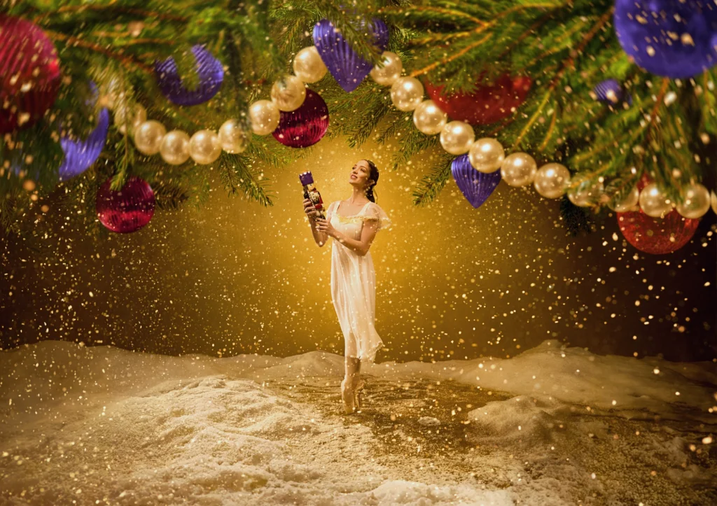 Clara stands in sous-sus on pointe, holding a Nutcracker doll, beneath the boughs of a decorated Christmas tree, where it seems to snow.