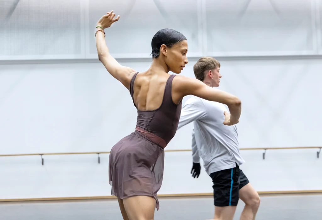 Leroy Mokgatle is shown in profile in a rehearsal studio. She twists upstage as she leans forward, arms curving in front of her and up and behind her. Her hair is slicked back against her skull; she wears a dusty purple leotard and a sheer ballet skirt in a slightly lighter shade.