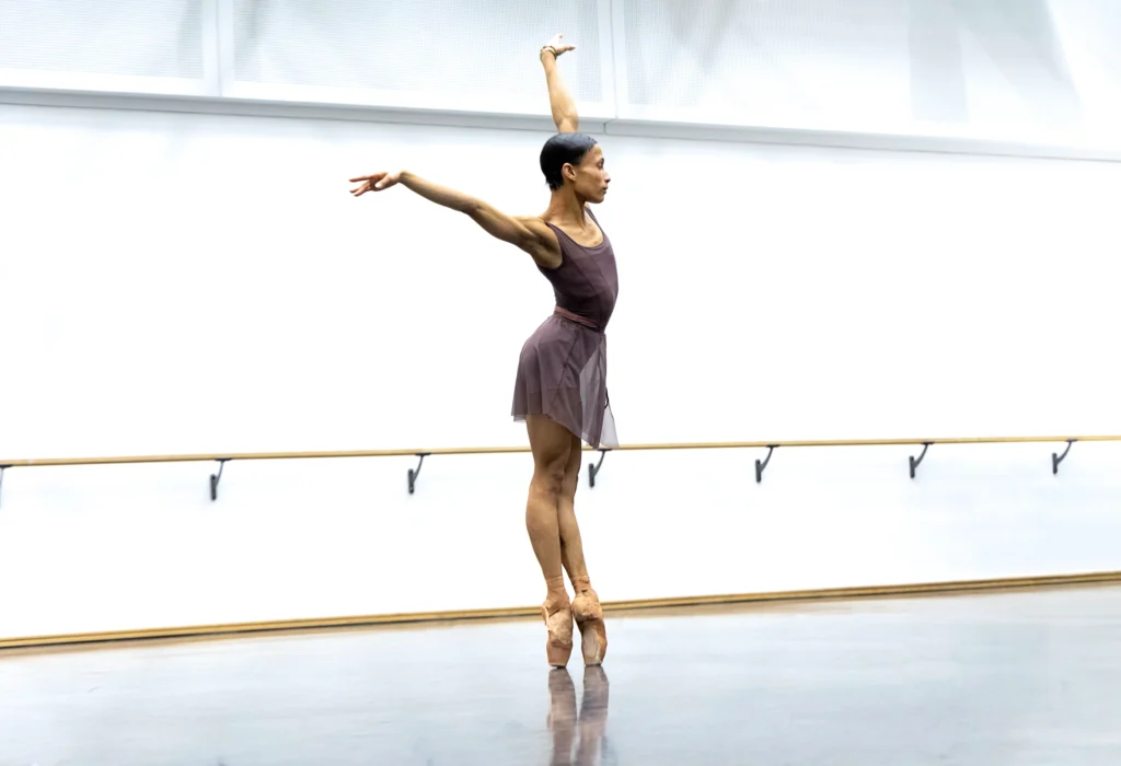 Leroy Mokgatle is show in profile as she poses in a sous-sus on pointe, arms in an exaggerated V overhead. She wears a dusty purples leotard, a sheer ballet skirt in a lighter shade, and pointe shoes pancaked to match her skin tone.