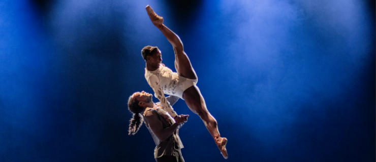 A male dancer lifting a female dancer as she tilts and pikes her body in the air.