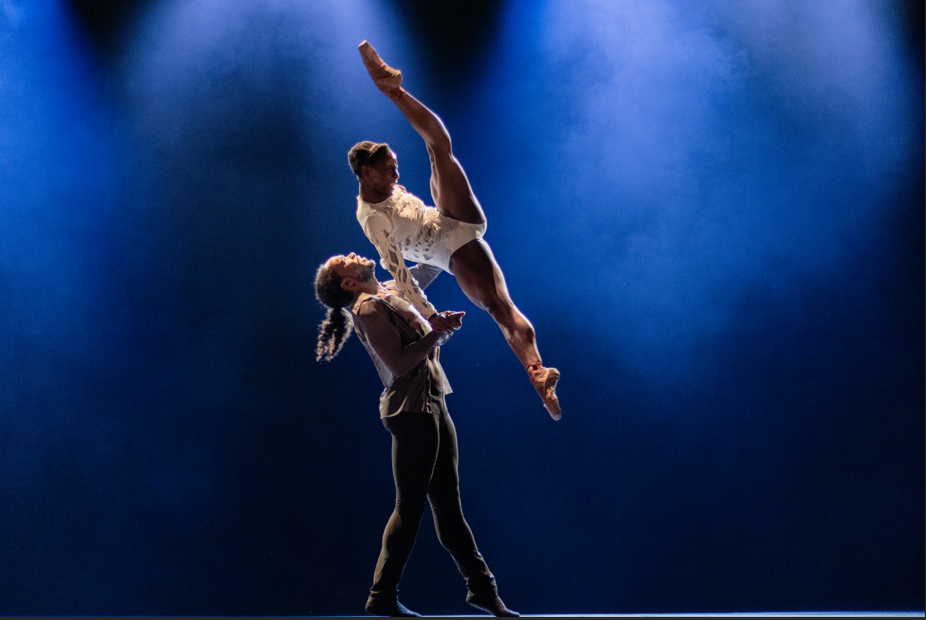 A male dancer lifting a female dancer as she tilts and pikes her body in the air.