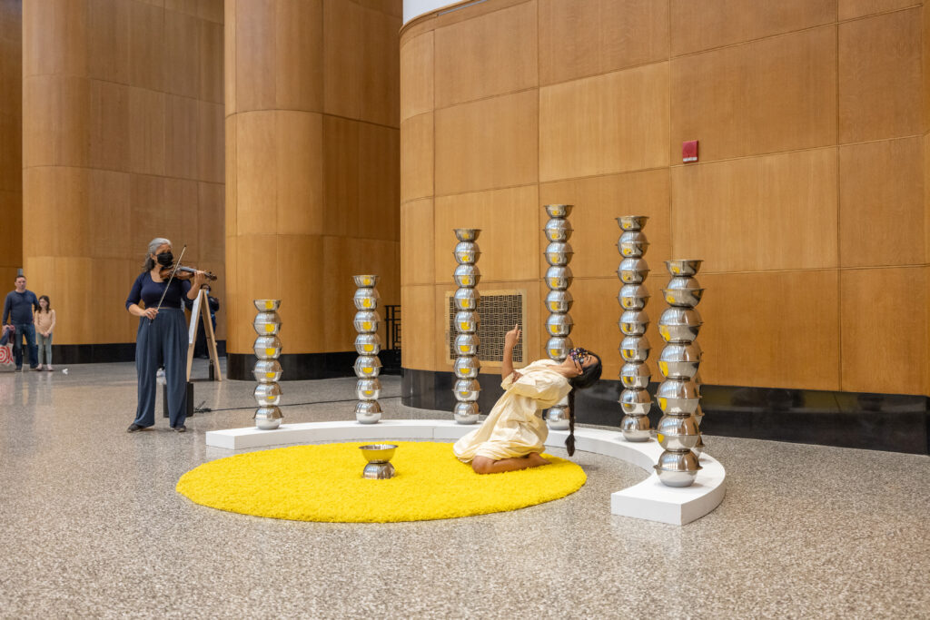 Smita Sen kneeling on a circular yellow rug as she performs along side a violinist. She is surrounded by a semi circle of metal towers.