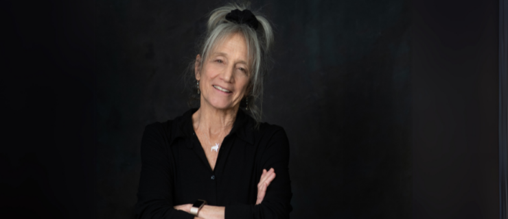 Liz Lerman's headshot. An woman with a gray ponytail standing with her arms crossed against a dark background