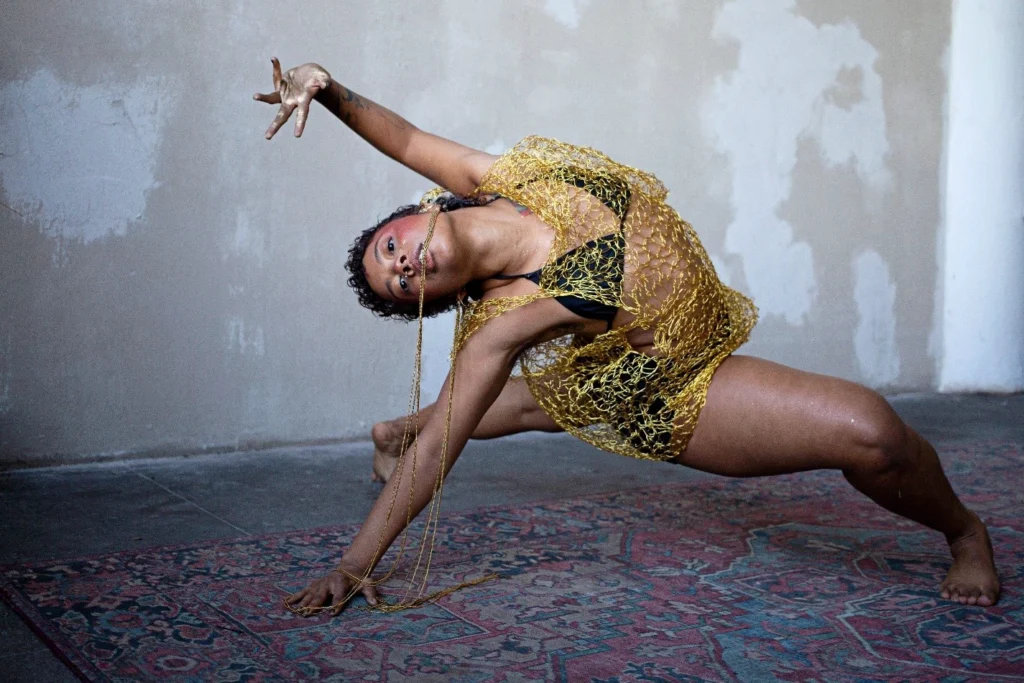 A dancer wearing gold netting over black briefs and bralette holds a long gold chain in their teeth. They lunge to the floor and arch back, supporting themself on one arm as their torso reaches parallel to the floor. They dance on a dusty patterned carpet, before a concrete wall splattered with white paint. 