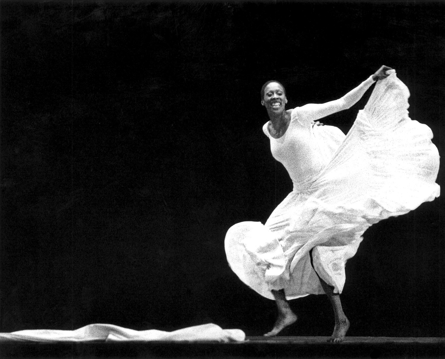 In a black and white archival image, Judith Jamison grins infectiously as she dances. She wears a long white dress, the skirt of which she lifts into a ripple with one arm as she moves lithely on the balls of her feet, knees bent.