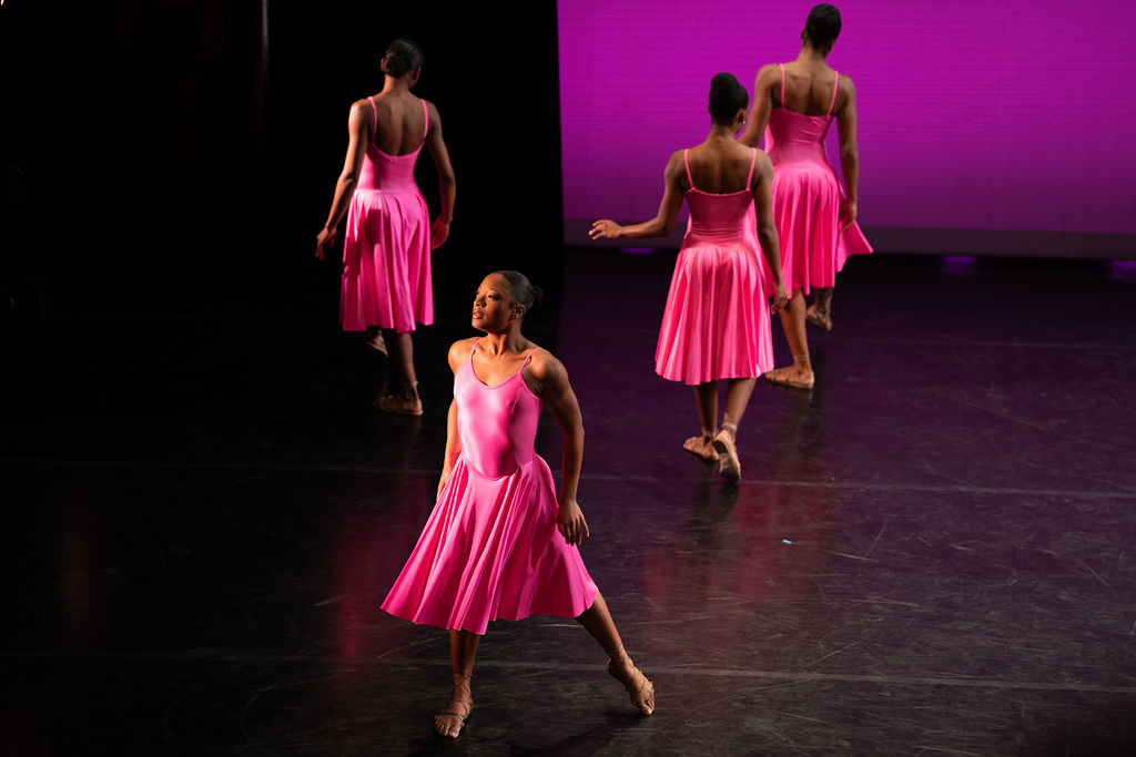 A dancer in a flowing pink dress looks wistfully offstage as her skirt whirls around her; her arms float just away from her sides as she inhales. Three dancers costumed in the same dress walk upstage, backs turned to the audience.