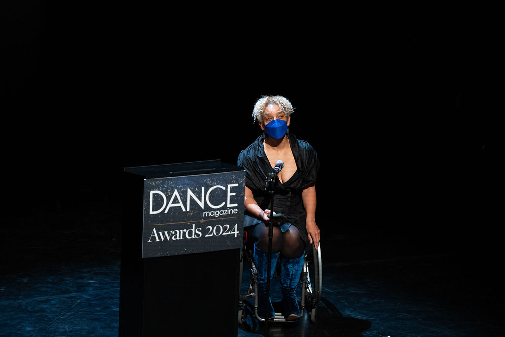 Alice Sheppard looks out at the audience from behind a microphone at the 2024 Dance Magazine Awards. She wears a KN95 match that is blue to match her boots; one hand rests on the wheel of her chair while the other holds her phone with her presentation notes.