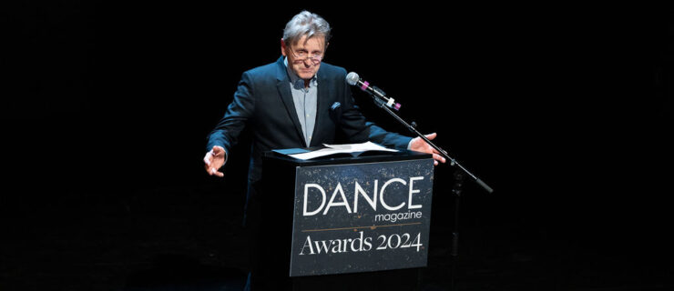 Mikhail Baryshnikov stands behind a podium that reads "Dance Magazine Awards 2024," his hands open to the audience as he speaks.