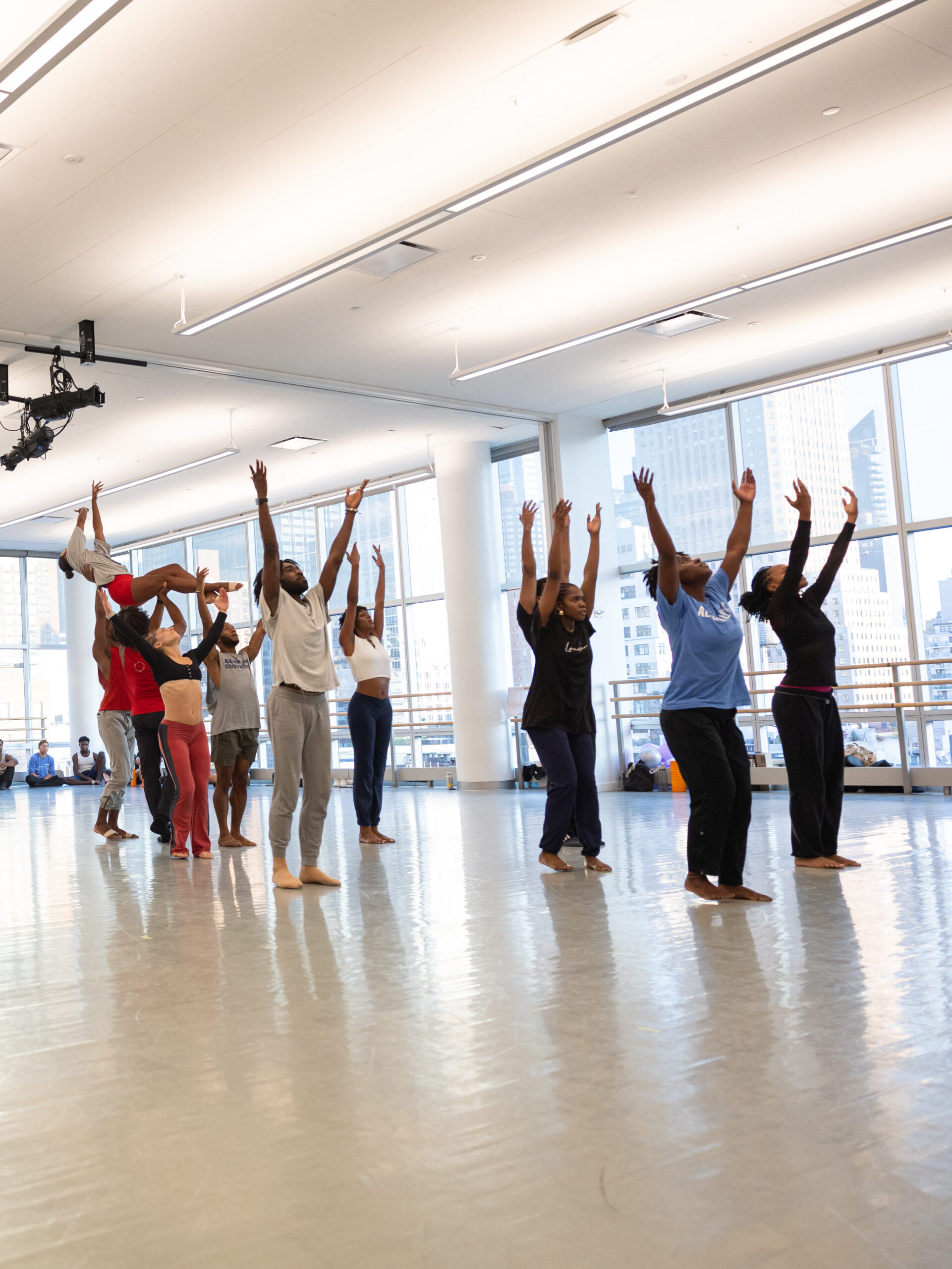 Ailey dancers lifting their arms up in rehearsal with a group of dancer lifting another dancer in the air behind them