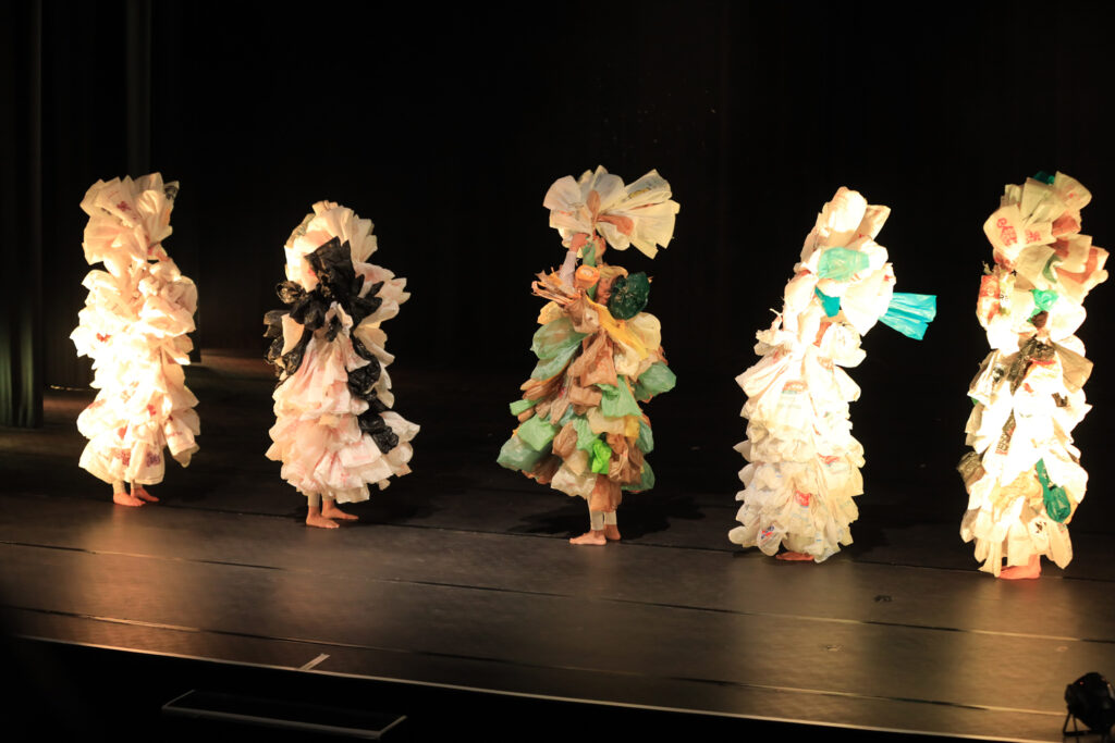 Five dancers stand in a line on a black stage, their bodies almost entirely obscured by their fantastical costumes made from hundreds of plastic bags.